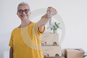 Senior happy woman holding keys of the new apartment standing in the empty home with moving boxes on the floor - concept of active