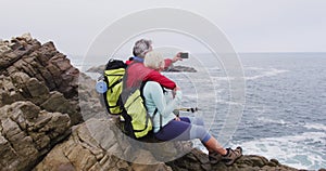 Senior hiker couple with backpacks sitting on the rocks and taking a selfie on smartphone