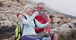 Senior hiker couple with backpacks sitting on the rocks and waving while having a video call