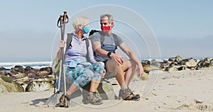 Senior hiker couple wearing face masks with backpacks and hiking poles talking sitting on rock