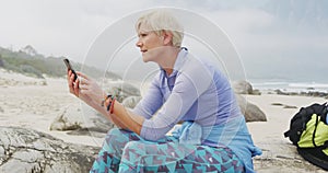 Senior hiker woman with backpack using smartphone while sitting on rock hiking on the beach.