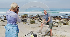 Senior hiker woman taking pictures of senior hiker man using digital camera on the beach.