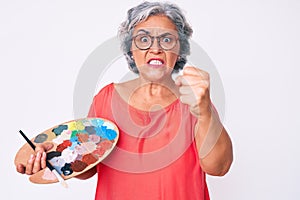 Senior hispanic grey- haired woman holding paintbrush and palette annoyed and frustrated shouting with anger, yelling crazy with