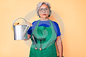 Senior hispanic grey- haired woman wearing gardener apron and gloves holding watering can thinking attitude and sober expression