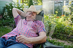 Senior hispanic man in hat sitting leaning back on chair sleeping in outdoor summer flower garden