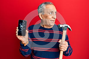 Senior hispanic man holding broken smartphone showing cracked screen and hammer smiling looking to the side and staring away