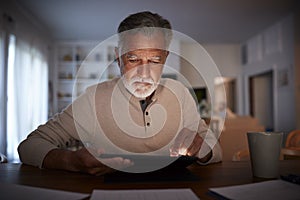 Senior Hispanic man sitting at a table reading an e book at home in the evening, close up