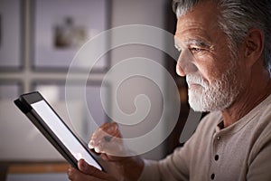 Senior Hispanic man sitting at a table using a tablet computer  in the evening, close up, side view