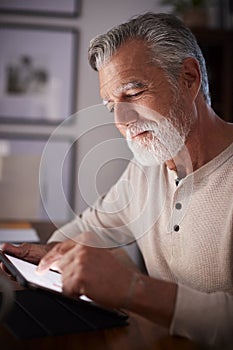 Senior Hispanic man sitting at a table using a tablet computer in the evening, close up, vertical