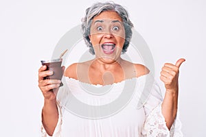 Senior hispanic woman drinking mate infusion pointing thumb up to the side smiling happy with open mouth