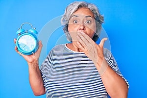 Senior hispanic woman holding alarm clock covering mouth with hand, shocked and afraid for mistake