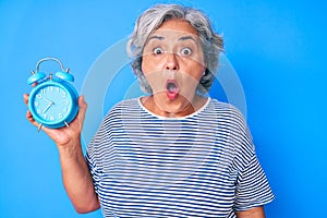 Senior hispanic woman holding alarm clock scared and amazed with open mouth for surprise, disbelief face