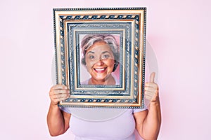 Senior hispanic woman holding empty frame smiling happy and positive, thumb up doing excellent and approval sign