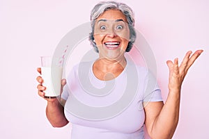 Senior hispanic woman holding glass of milk celebrating victory with happy smile and winner expression with raised hands