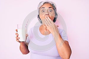 Senior hispanic woman holding glass of milk covering mouth with hand, shocked and afraid for mistake