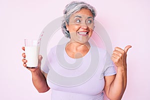 Senior hispanic woman holding glass of milk pointing thumb up to the side smiling happy with open mouth