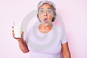 Senior hispanic woman holding glass of milk scared and amazed with open mouth for surprise, disbelief face