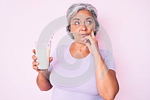 Senior hispanic woman holding glass of milk serious face thinking about question with hand on chin, thoughtful about confusing