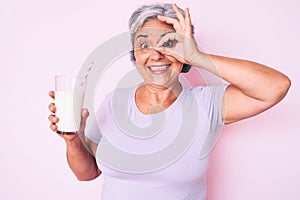 Senior hispanic woman holding glass of milk smiling happy doing ok sign with hand on eye looking through fingers