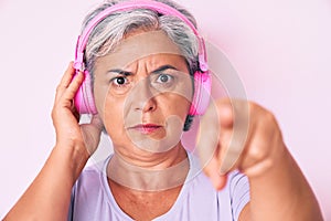 Senior hispanic woman listening to music using headphones pointing with finger to the camera and to you, confident gesture looking