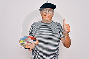 Senior hoary artist man wearing french beret drawing using paintbrush and palette with colors happy with big smile doing ok sign,