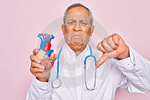 Senior hoary doctor man wearing stethoscope holding plastic heart over pink background with angry face, negative sign showing