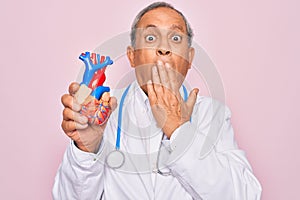 Senior hoary doctor man wearing stethoscope holding plastic heart over pink background cover mouth with hand shocked with shame