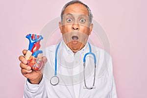 Senior hoary doctor man wearing stethoscope holding plastic heart over pink background scared in shock with a surprise face,