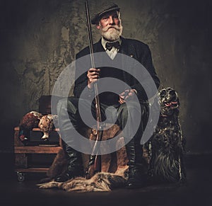 Senior hunter with a english setter and shotgun in a traditional shooting clothing, sitting on a dark background.