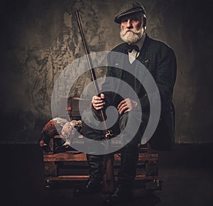 Senior hunter with a shotgun and pheasants in a traditional shooting clothing, sitting on a dark background.