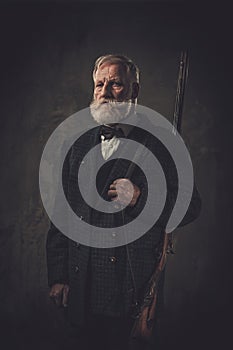 Senior hunter with a shotgun in a traditional shooting clothing, posing on a dark background.