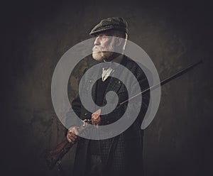 Senior hunter with a shotgun in a traditional shooting clothing, posing on a dark background.