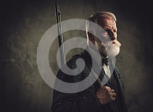 Senior hunter with a shotgun in a traditional shooting clothing, posing on a dark background.