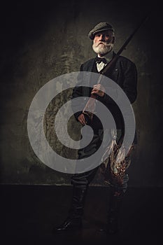 Senior hunter with a shotgun in a traditional shooting clothing, posing on a dark background.