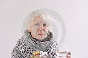 Senior ill woman with glass of water and peels in scarf freezing on white background.