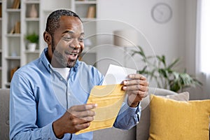 Senior joyful man sitting on sofa at home, satisfied smiling african american man reading mail message holding letter