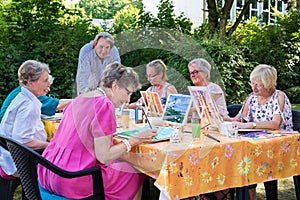 Senior ladies taking art lessons sitting outdoors at the table and painting pictures, following male instructors