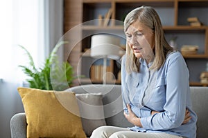Senior lady holding stomach with both arms while suffering from severe spasms and abdominal pain indoors. Frowning woman