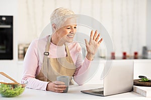 Senior Lady Making Video Call On Laptop Sitting In Kitchen