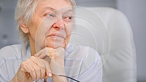 Senior lady pensioner holds head on wrinkly hands with glasses and looks aside with smile sitting on chair at home