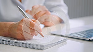 Senior lady teacher with wrinkly hands holds coloured pen over white page of paper notebook near grey laptop on table