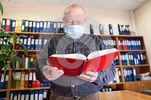 Senior male archivist holding open red notebook in hands, looking at camera, man wearing face mask due Covid-19 pandemic