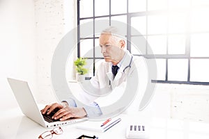 Senior male doctor sitting behind his laptop and working in doctor`s office photo