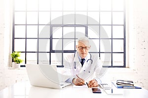 Senior male doctor sitting at desk in doctor`s room and working