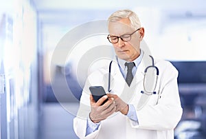 Senior male doctor text messaging on mobile phone while standing in doctor`s office photo