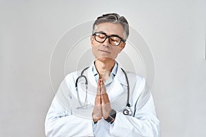 Senior male doctor wearing white coat and glasses, with stethoscope around neck keeping eyes closed and praying for help