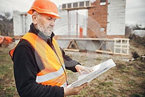 Senior male foreman in hardhat checking blueprints of building new modern house. Man engineer or construction worker looking at