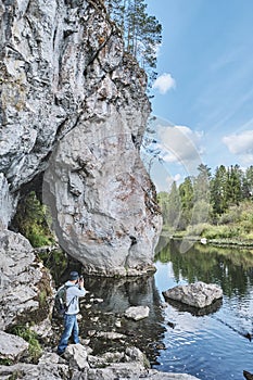 Senior male hiker taking pictures on mobile phone on bank of river near rock Summer nature landscape