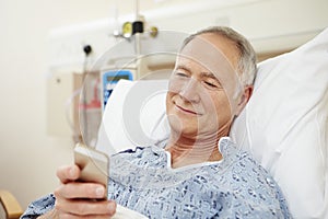 Senior Male Patient Using Mobile Phone In Hospital Bed