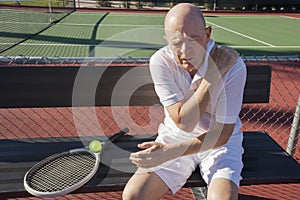 Senior male tennis player with shoulder pain sitting on bench at court